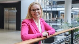A person with long blond hair, wearing glasses and a bright pink blazer, stands smiling by a railing in a modern indoor office setting.