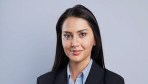 A woman with long dark hair wearing a black blazer and light blue collared shirt poses in front of a plain light gray background.