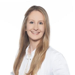 A woman with long blonde hair wearing a white lab coat smiles at the camera against a plain white background.