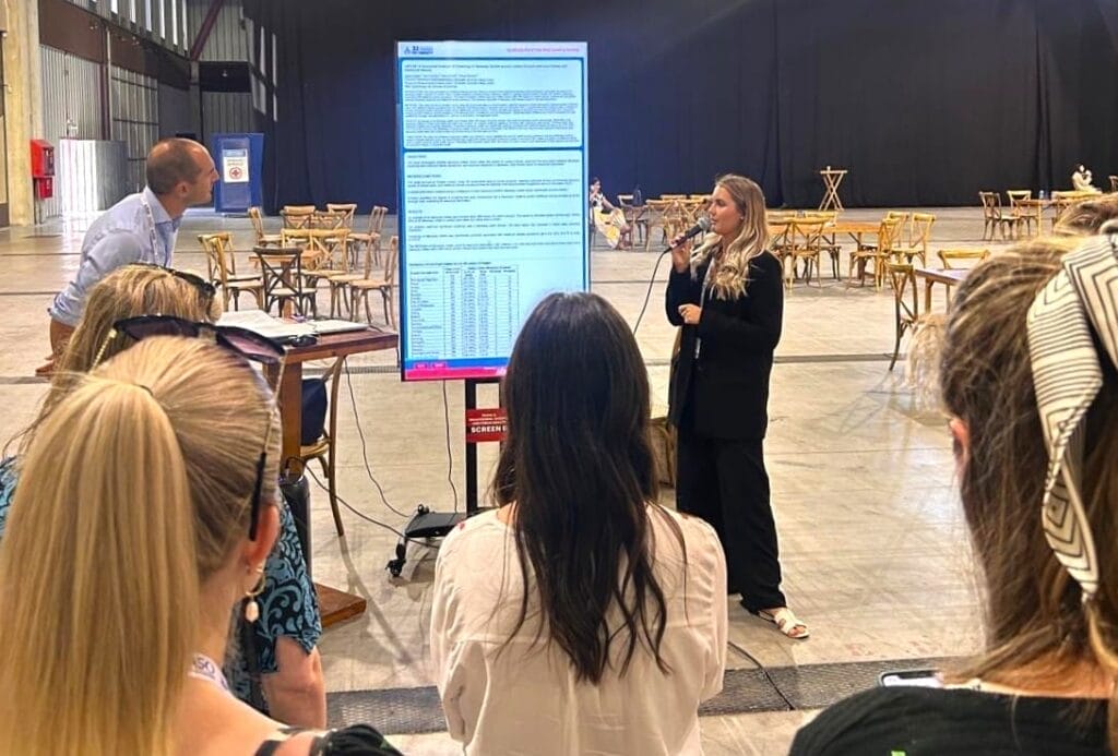 A woman speaks into a microphone while presenting data on a large screen to a small audience in a spacious indoor venue with tables and chairs.