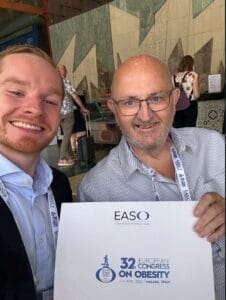 Two men smiling and holding a sign for the 32nd European Congress on Obesity, Málaga 2025. Conference attendees are visible in the background.