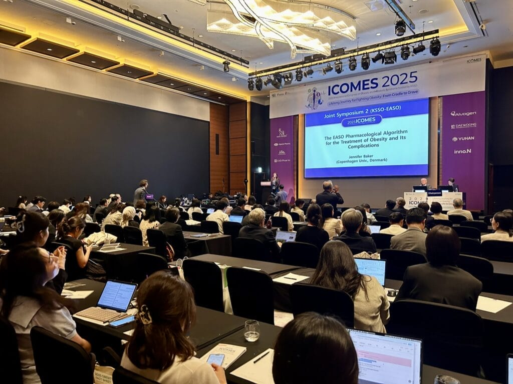A conference room filled with attendees watching a presentation on obesity treatment algorithms at ICOMES 2025; several people are taking notes on laptops.