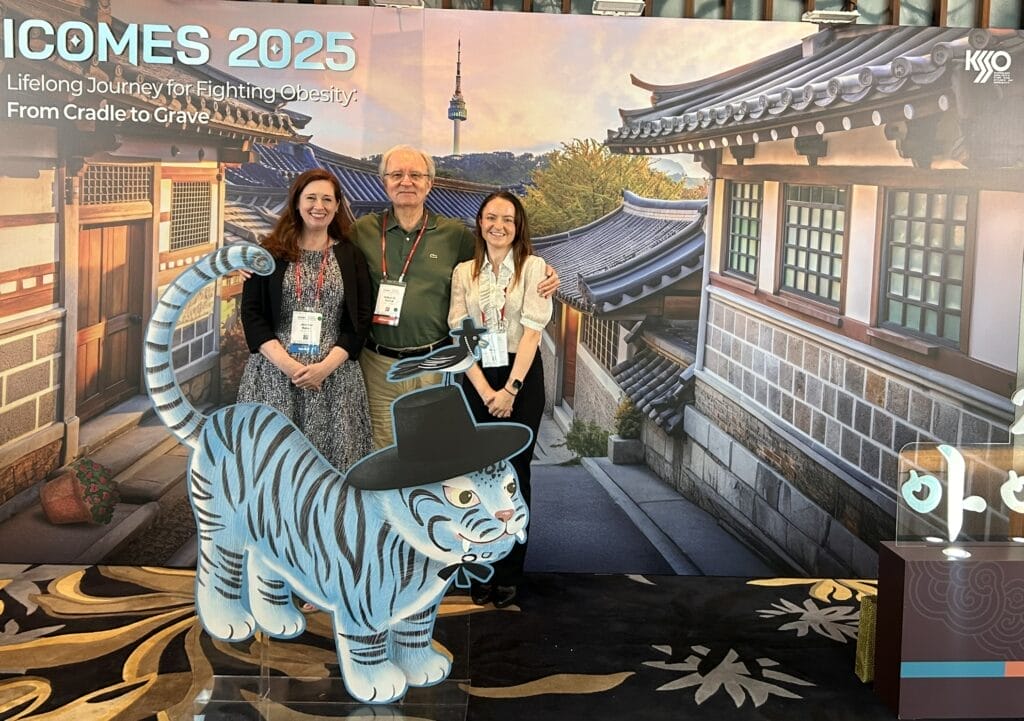 Three people stand indoors posing behind a blue tiger cutout at a medical conference with a traditional Korean street backdrop and a sign reading "ICOMES 2025: Lifelong Journey for Fighting Obesity.