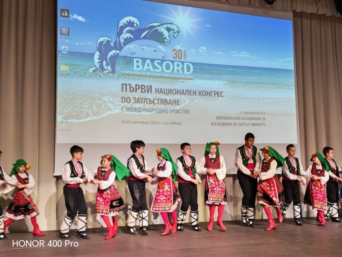 A group of children in traditional Bulgarian folk costumes dance on stage in front of a large conference screen with Bulgarian and English text.